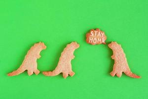 biscoitos caseiros em formas de dinossauros com inscrição - fique em casa - sobre fundo verde, vista superior. biscoito doce com esmalte branco. conceito de distanciamento social. foto