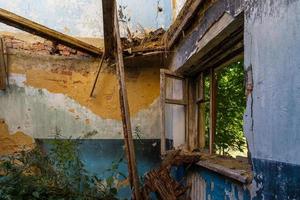 janela de madeira quebrada e sala com grama alta, vista dentro de um dormitório meio destruído abandonado à luz do dia de verão foto