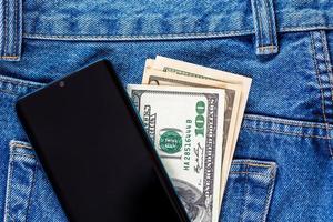 notas de dólar americano e smartphone sobre o bolso traseiro esquerdo da calça jeans. foto