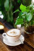 xícara de cappuccino quente e vaso de vidro com planta verde na mesa de madeira no café. foto