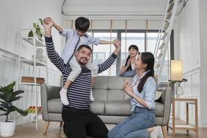 família tailandesa asiática junto, papai brinca e brinca com crianças e mamãe carregando e segurando o filho nos ombros na sala de estar em casa, momentos de lazer felizes, fim de semana adorável, estilo de vida doméstico de bem-estar. foto