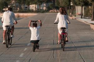 família feliz desfrutando de uma bela manhã à beira-mar juntos, pais andando de bicicleta e seu filho andando de scooter elétrico. foco seletivo foto