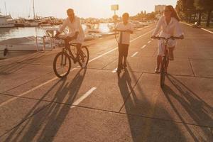 família feliz desfrutando de uma bela manhã à beira-mar juntos, pais andando de bicicleta e seu filho andando de scooter elétrico. foco seletivo foto