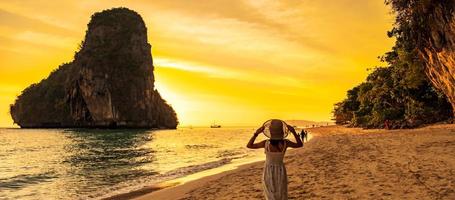 turista de mulher de vestido branco e chapéu andando na praia da caverna de phra nang ao pôr do sol, railay, krabi, tailândia. férias, viagens, verão, desejo de viajar e conceito de férias foto