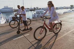 família feliz desfrutando de uma bela manhã à beira-mar juntos, pais andando de bicicleta e seu filho andando de scooter elétrico. foco seletivo foto