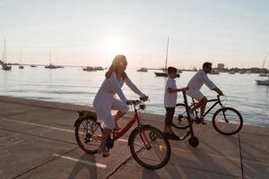 família feliz desfrutando de uma bela manhã à beira-mar juntos, pais andando de bicicleta e seu filho andando de scooter elétrico. foco seletivo foto