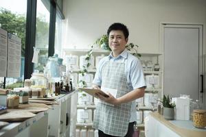 retrato de um lojista asiático sorrindo e olhando para a câmera, organiza produtos naturais na loja de refil, mercearia com desperdício zero e estilos de vida sustentáveis, ecologicamente corretos e sem plástico. foto