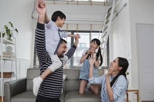 família tailandesa asiática junto, papai brinca e brinca com crianças e mamãe carregando e segurando o filho nos ombros na sala de estar em casa, momentos de lazer felizes, fim de semana adorável, estilo de vida doméstico de bem-estar. foto