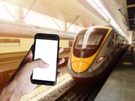 mão de homem segurando smartphone preto com tela em branco na frente do trem moderno indo na estação ferroviária. espaço para texto ou design foto