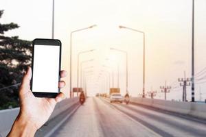 mão de adulto segurando smartphone preto com tela em branco atrás do carro e moto na estrada com desfoque de movimento de fundo. espaço para texto ou design foto