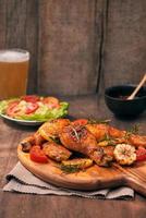 pernas de frango grelhado assado na grelha na tábua de madeira com molho de tomate em uma tigela, tomate fresco e folhas de alface, pimenta amarga, caneca de vidro de cerveja foto