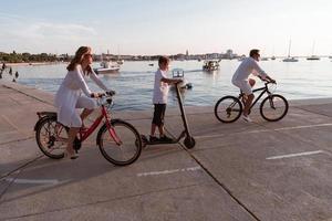 família feliz desfrutando de uma bela manhã à beira-mar juntos, pais andando de bicicleta e seu filho andando de scooter elétrico. foco seletivo foto