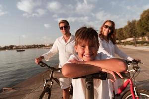 família feliz desfrutando de uma bela manhã à beira-mar juntos, pais andando de bicicleta e seu filho andando de scooter elétrico. foco seletivo foto