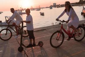 família feliz desfrutando de uma bela manhã à beira-mar juntos, pais andando de bicicleta e seu filho andando de scooter elétrico. foco seletivo foto