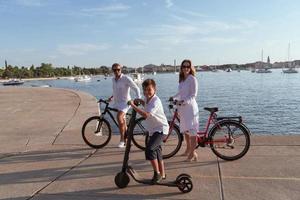 família feliz desfrutando de uma bela manhã à beira-mar juntos, pais andando de bicicleta e seu filho andando de scooter elétrico. foco seletivo foto