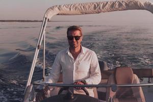 um determinado empresário sênior em roupas casuais e óculos de sol aproveita suas férias dirigindo um barco de luxo ao pôr do sol. foco seletivo foto
