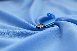 cartão de memória flash usb azul brilhante com um laço azul encontra-se em um cobertor de tecido de lã azul claro macio e peludo com muitas dobras em relevo. dispositivo de armazenamento de memória em design feminino foto