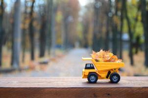 um pequeno caminhão amarelo de brinquedo é carregado com folhas amarelas caídas. o carro fica em uma superfície de madeira contra um fundo de um parque de outono embaçado. limpeza e remoção de folhas caídas. trabalhos sazonais foto