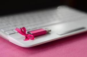 cartão de memória flash usb rosa brilhante com um laço rosa encontra-se em um cobertor de tecido de lã rosa claro macio e peludo ao lado de um laptop branco. design de presente feminino clássico para um cartão de memória foto