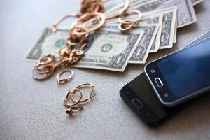 muitos anéis, brincos e colares dourados caros com grande quantidade de notas de dólar perto de smartphones. loja de penhores foto