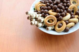 túbulos crocantes, bolas de chocolate derretendo e bagels estão em um prato branco sobre uma mesa de madeira. mistura de vários doces foto