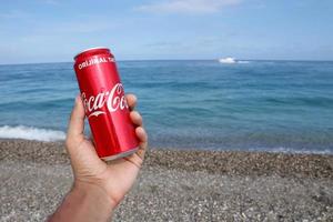 antalya, turquia - 18 de maio de 2021 lata vermelha original da coca cola encontra-se em pequenas pedras redondas perto da costa do mar. lata de coca-cola na mão masculina na praia foto