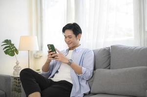 jovem asiático usando telefone inteligente no sofá na sala de estar foto