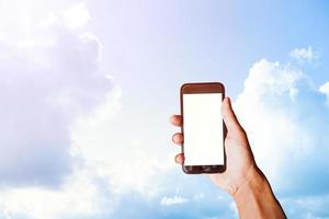 mão de homem segurando smartphone preto com tela em branco atrás de nuvens brancas com fundo de céu azul em um dia brilhante. espaço para texto ou design foto