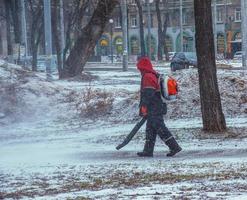 trabalho sazonal de serviços públicos da cidade no parque. um trabalhador com um soprador de mochila motorizado sopra neve de um caminho do parque foto