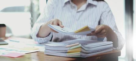 documentos de negócios conceito empregado mulher mãos trabalhando em pilhas de arquivos de papel para pesquisar e verificar documentos inacabados alcança em papéis de pastas no escritório de mesa de trabalho ocupado. foco suave foto