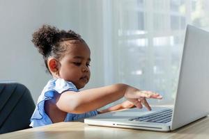 pequeno americano fofo usa laptop para estudar em casa, fazer lição de casa, senta-se em uma mesa de madeira na sala de estar. foto