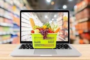 corredor de supermercado fundo desfocado com computador portátil e cesta de compras no conceito on-line de mercearia de mesa de madeira foto