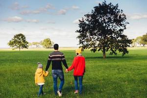 três membros amigáveis da família caminham juntos no prado verde, mantêm as mãos, têm uma conversa agradável, desfrutam da união e das belas paisagens, olham para longe, ficam de costas para a câmera foto