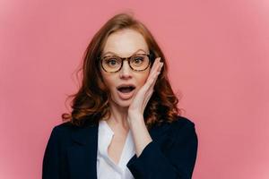 realmente de jeito nenhum a mulher ruiva chocada toca a bochecha, abre a boca amplamente, olha surpreendentemente para a câmera, descobre notícias chocantes, usa óculos, terno formal elegante, isolado no fundo rosa foto