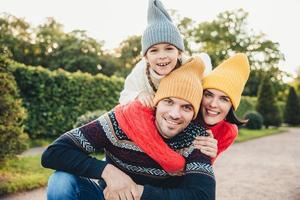 retrato ao ar livre de homem bonito dar carona para sua esposa e filha, usar roupas quentes, ter expressões felizes, apoiar uns aos outros. família abraça ao ar livre, sorri agradavelmente para a câmera foto