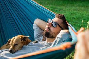 sorrindo atraente homem europeu em óculos de sol está descansando na rede com seu cachorrinho fofo. foto