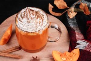 café com leite de abóbora com chantilly em fundo escuro. bebida quente de outono com canela e especiarias em caneca de vidro em uma bandeja de madeira. foto