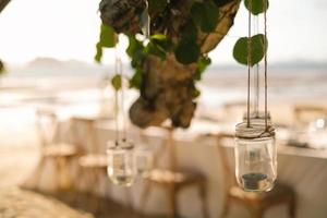 feche a vela do frasco pendurada na árvore com a configuração do jantar de casamento de mesa longa na praia na tailândia à noite. conceito de festa de casamento. decoração restaurante ao ar livre na praia. foto