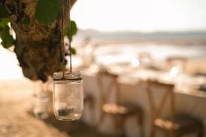 feche a vela do frasco pendurada na árvore com a configuração do jantar de casamento de mesa longa na praia na tailândia à noite. conceito de festa de casamento. decoração restaurante ao ar livre na praia. foto