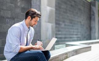 jovem homem caucasiano sentado e trabalhando em projeto freelance usando computador portátil no escritório externo. foto