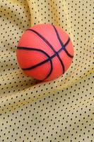 pequena bola de basquete de borracha laranja encontra-se em uma textura de tecido de roupa de camisa esportiva amarela e fundo com muitas dobras foto