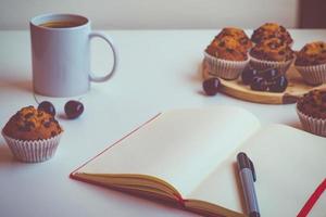 um caderno vazio com uma capa vermelha sobre uma mesa ao lado de uma caneta, uma xícara de café, muffins de chocolate e algumas cerejas foto