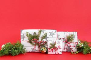 caixas de presente de natal embrulhadas em papel branco-cinza com fitas vermelho-brancas sobre fundo vermelho, decoradas com galhos de abeto e decorações de natal verdes brilhantes. conceito de natal e ano novo. foto