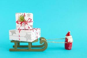 papai noel está carregando trenó de madeira verde com duas caixas de presente embrulhadas de papel branco e cinza com fita vermelho-branca, decorada de galho de abeto em um fundo azul brilhante. foto