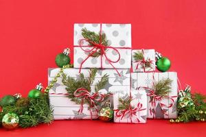 caixas de presente de natal embrulhadas em papel branco-cinza com fitas vermelho-brancas sobre fundo vermelho, decoradas com galhos de abeto e decorações de natal verdes brilhantes. conceito de natal e ano novo. foto