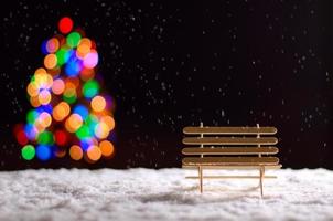 banco de madeira ao nevar no chão na temporada de inverno e luzes coloridas de bokeh da árvore de natal. foto