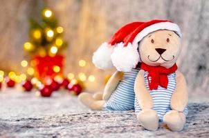 boneca de ursinho de pelúcia com chapéu de Papai Noel e cachecol com bokeh luz de fundo da árvore de natal. foto