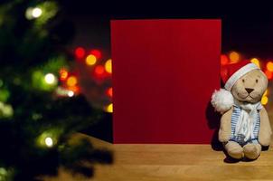 ursinho de pelúcia do papai noel sentado ao lado do cartão vermelho vazio com árvore de natal e fundo colorido de luzes bokeh. foto