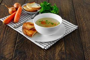 caldo fresco em uma tigela branca com ervas picadas com salsa e alho em uma mesa de madeira. sopa quente feita de carne de frango, cenoura, cebola, alho, salsa e pimenta. foto