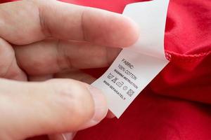 Segure a mão e leia no rótulo de roupas de instruções de lavagem de cuidados com a roupa branca na camisa de algodão vermelha foto